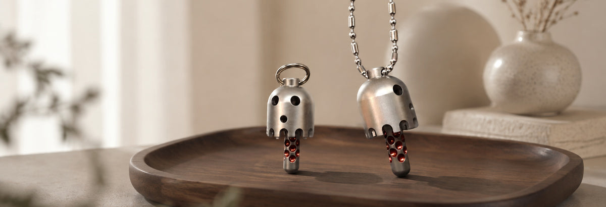 Two Daze bell pendants on a wooden tray, one with a ring and the other with a chain, beside a vase.